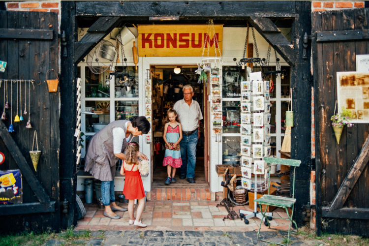 Ein Mann und ein Kind kommen aus dem Dorfladen, eine Frau und ein Kind stehen davor und schauen sich Ware an.