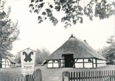 Eine alte Aufnahme in schwarz-weiß, die den Eingang zum Denkmalhof zeigt. Im Vordergrund ein Schild mit den Öffnungszeiten des Denkmalhofes, im Hintergrund das Bauernhaus und die Scheune Klockenhagen.