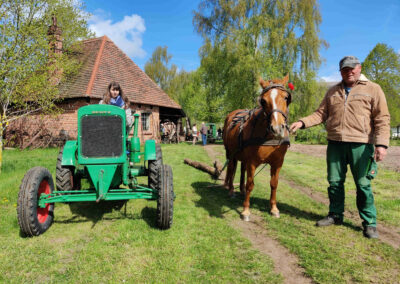 Ein Mädchen sitzt auf einen Traktor der Marke Deutz, daneben steht ein Pferd, das einen Baumstamm zieht.