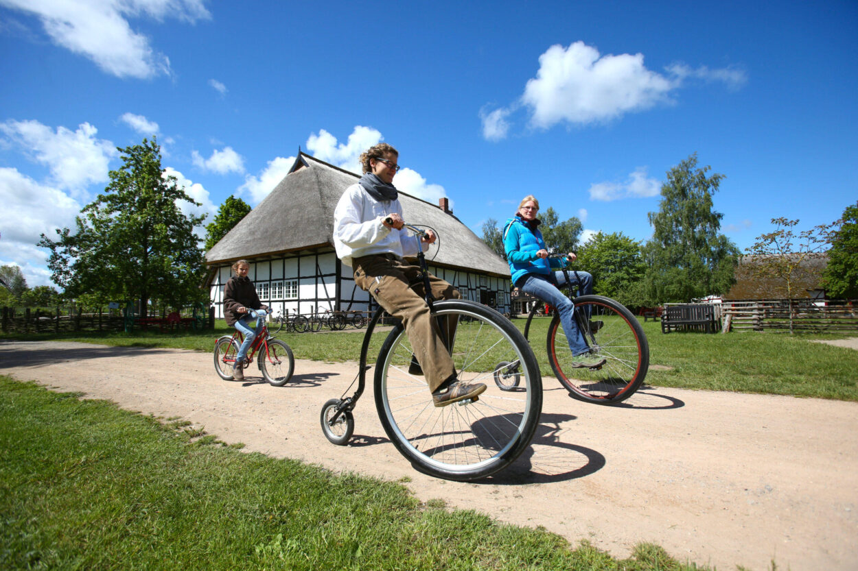 2 Frauen fahren Hochrad auf dem Gelände des Freilichtmuseums Klockenhagen, dahinter fährt ein Kind auf dem Fahrrad, im Hintergrund ist ein Bauernhaus zu sehen