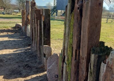 Ein aus Holzbalken und Feldsteinen gebauter Zaun zur Begrenzung des Gartens, im Hintergrund die Bockwindmühle.