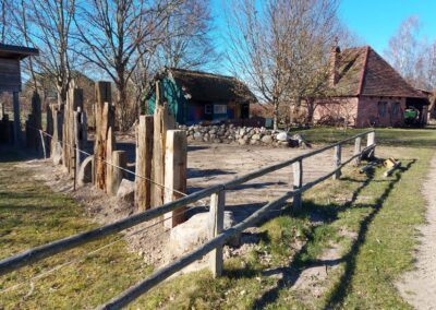 Ein aus Holzbalken und Feldsteinen gebauter Zaun zur Begrenzung des Gartens, im Hintergrund die Schmiede.
