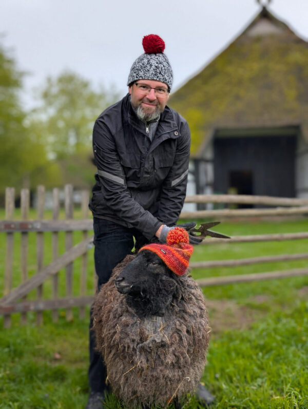 Ein Mann mit einer Wollmütze auf dem Kopf steht vor einem Schaf, das auch eine Wollmütze trägt. Der Mann hält eine Schere in der Hand.