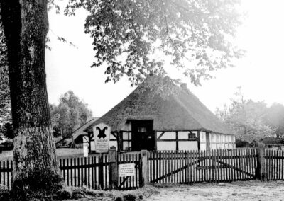Ein altes Schwarz-Weiß-Bild mit einem Bauernhaus und einem Zaun davor. Auf einem Schild steht "Denkmalhof".