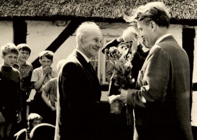 Ein altes Schwarz-Weiß-Bild. Ein Mann schüttelt einem älteren Mann die Hand und überreicht einen Blumenstrauß. Im Hintergrund sind weitere Menschen, vor allem Kinder, zu sehen.