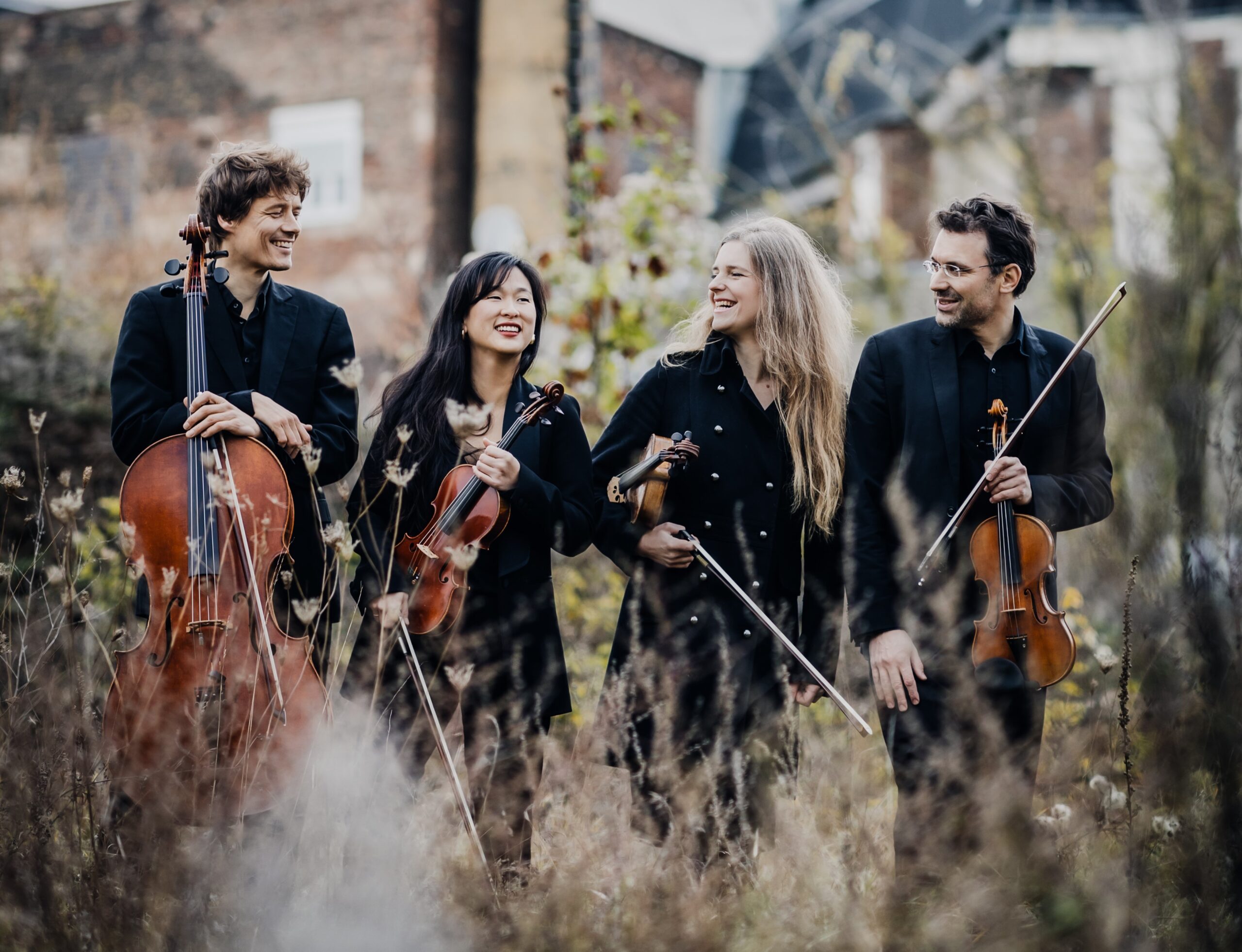 Ein Streichquartett, bestehend aus zwei Männern und zwei Frauen, die jeder ein Streichinstrument in der Hand halten, stehen vor einem Gebäude